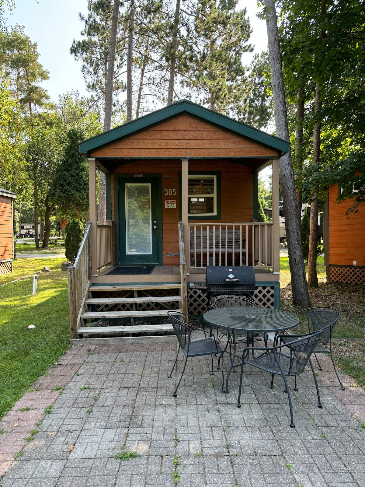 A charming cabin is presented with wooden siding and a welcoming front porch. A small table with four chairs is positioned on a brick patio. Surrounding trees provide natural shade, enhancing the serene outdoor setting.