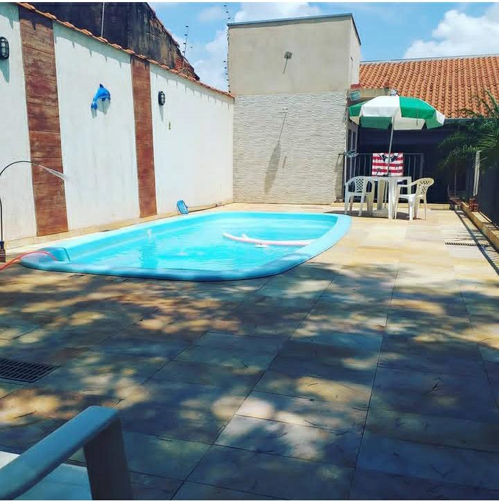 A private pool is situated in a sunlit courtyard, surrounded by smooth tiled flooring. A shaded seating area with chairs and an umbrella adds to the outdoor space, while adjacent walls provide privacy. Lush greenery is visible, enhancing the serene environment.
