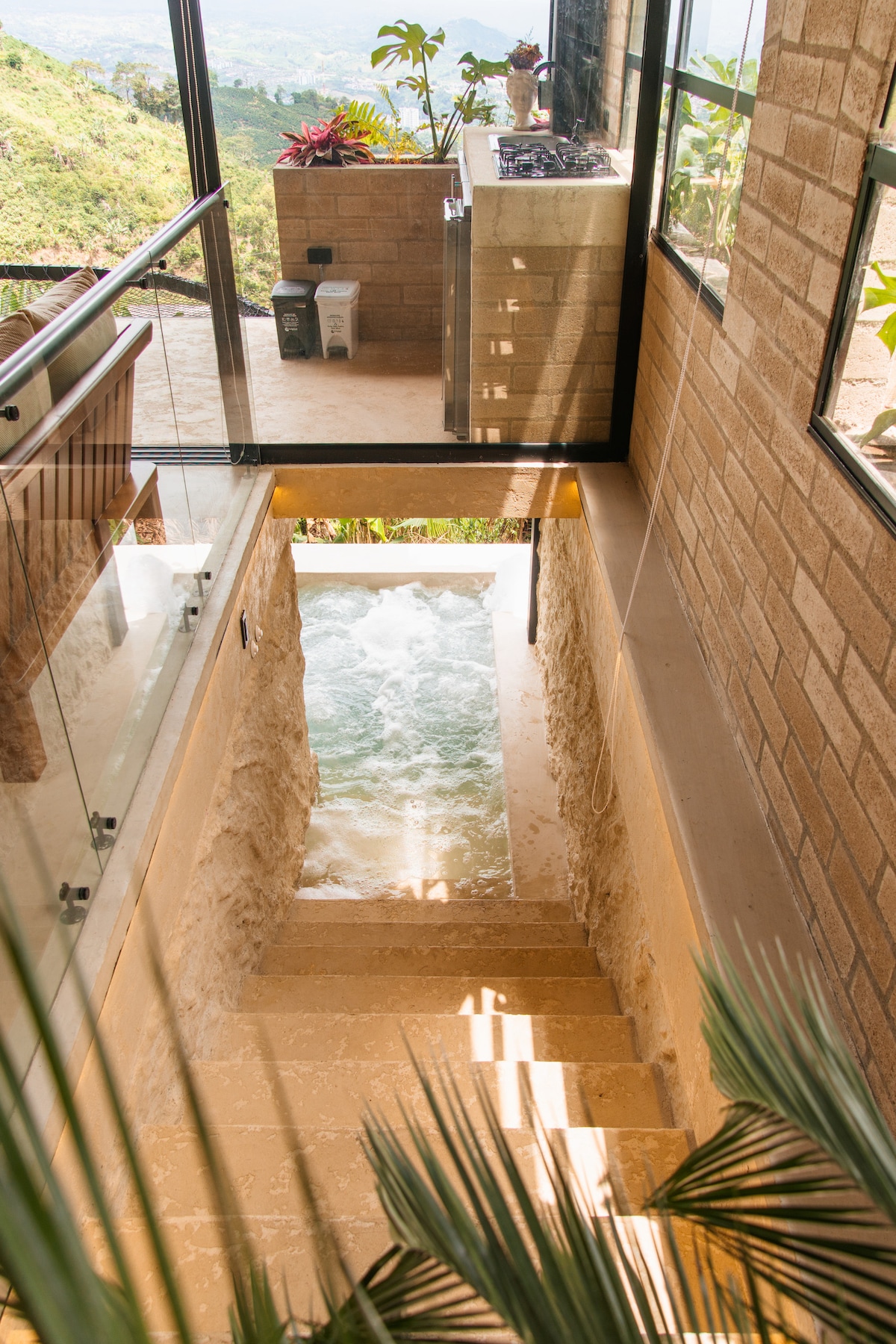 A staircase descends to a spacious indoor pool, with water cascading gently at the bottom. Natural light filters through large windows, illuminating the stone walls. Lush greenery is visible outside, creating a serene connection to the surrounding landscape.