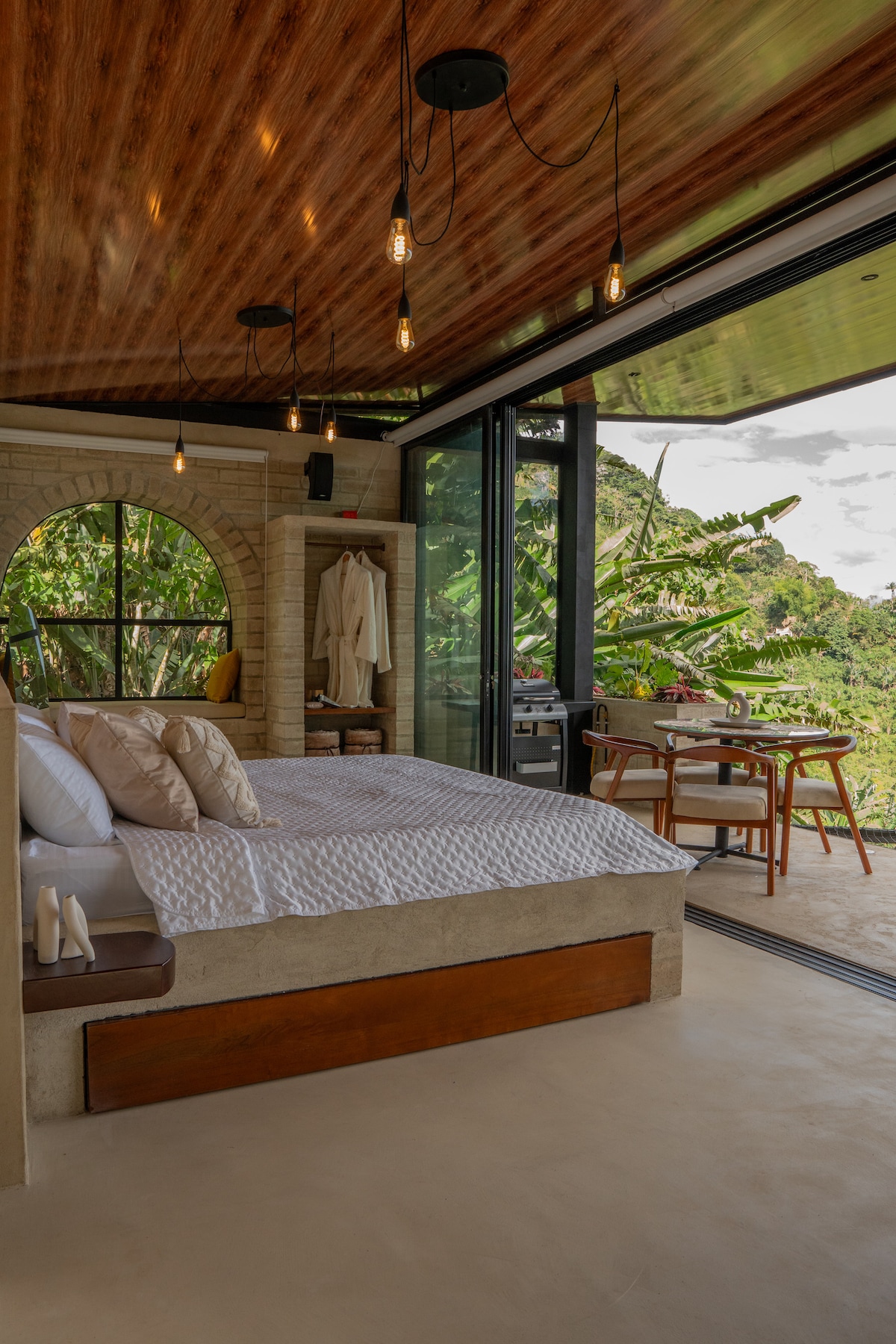 A bright and airy bedroom features a spacious bed with light-colored bedding, complemented by decorative pillows. Large glass doors open to a private outdoor seating area, surrounded by lush greenery. The ceiling showcases wooden beams, enhancing the natural ambiance.