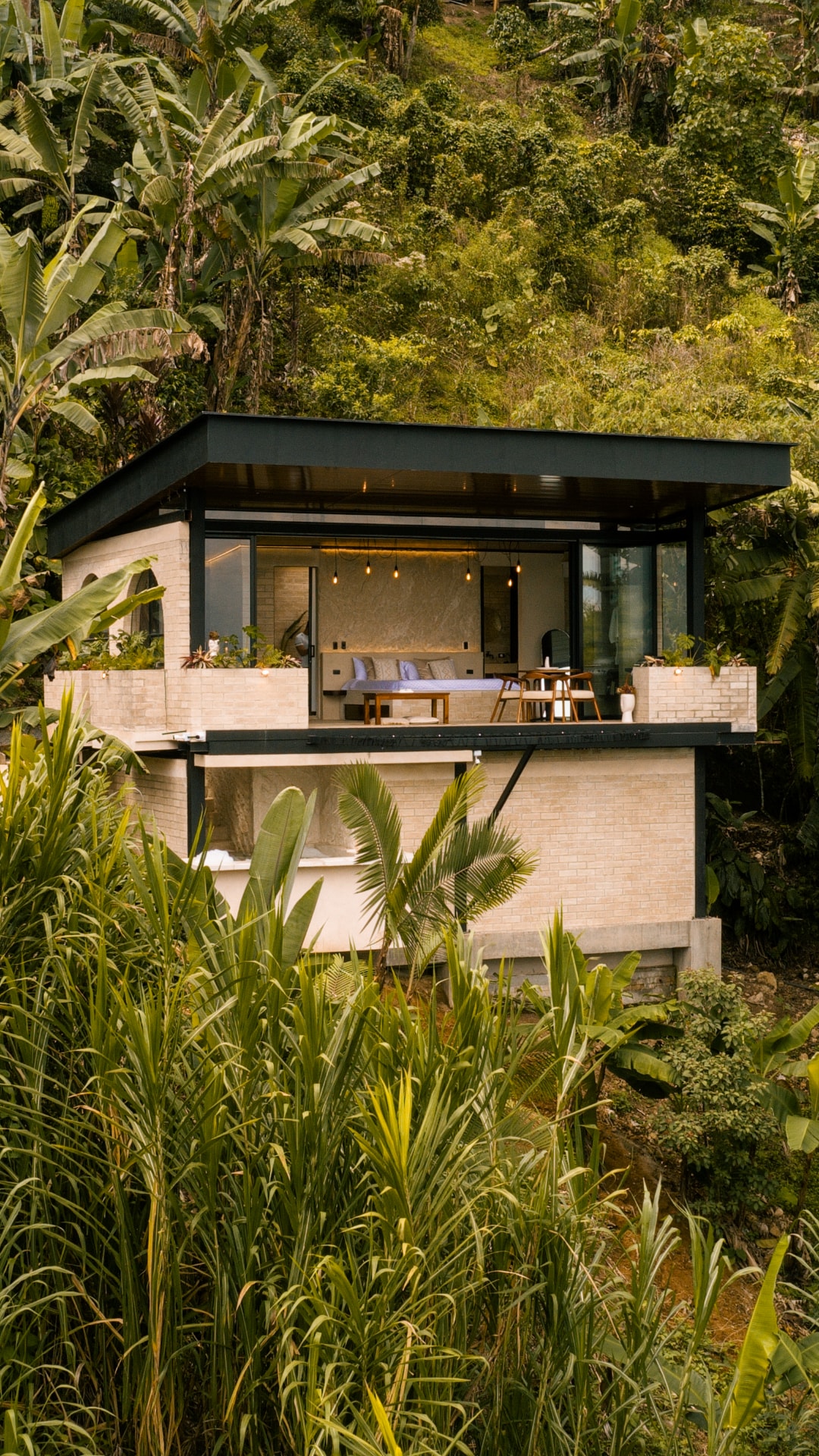 The modern cabin is surrounded by lush greenery, with large glass windows providing a view of the interior. Potted plants are placed on the balcony, enhancing the connection to nature. The structure's contemporary design contrasts with the vibrant landscape.