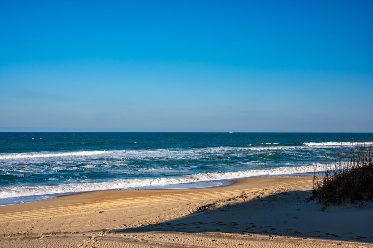 The image captures a serene beach scene featuring gentle waves lapping at the shore. A sandy beach stretches along the coastline beneath a clear blue sky, while sparse grasses are visible in the foreground. The tranquil atmosphere invites relaxation and beachfront enjoyment.