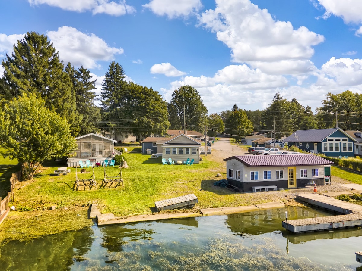 A serene aerial view of waterfront cottages surrounded by green lawns and tall trees. A dock extends into the calm water, with two distinct cottages visible along the shore. Colorful seating areas are noted on the lawn, offering space for outdoor relaxation.