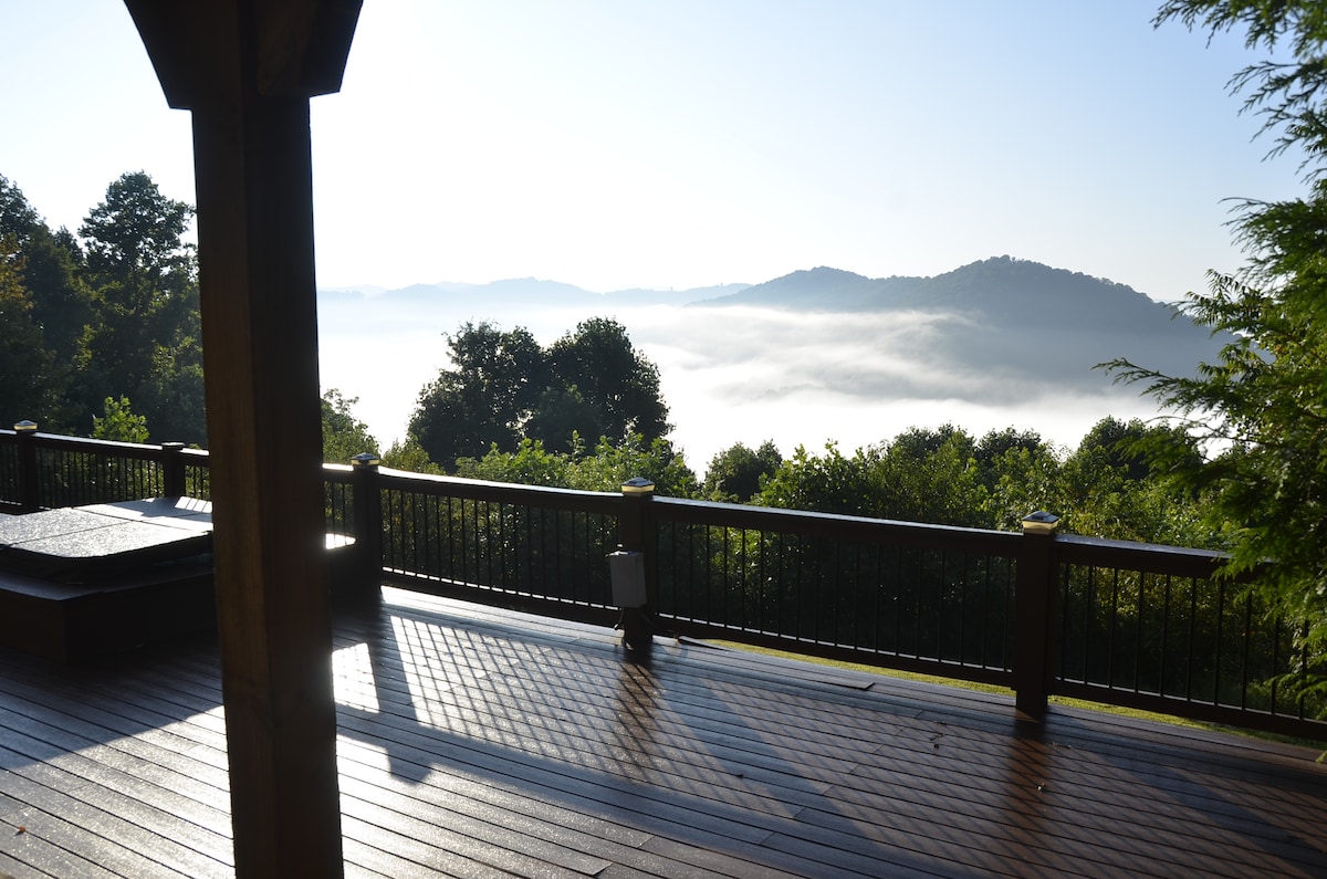 An expansive wooden deck offers panoramic views of mist-covered mountains and valleys. The sun illuminates the surface, highlighting the natural wood grain. A sunken hot tub is visible to the left, creating a tranquil outdoor space for relaxation.