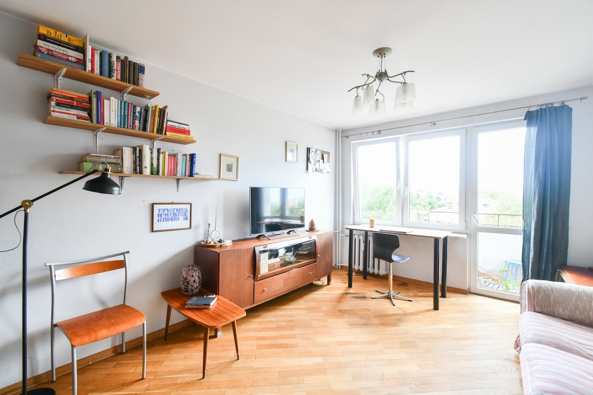 A minimalist living area features a comfortable seating space and a flat-screen television. Light wooden flooring complements the warm tones of the furniture. A wall-mounted bookshelf holds various books, while a small table and workstation are positioned near the large windows allowing ample natural light.