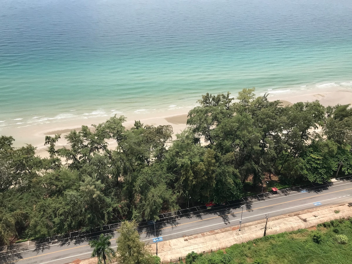 A panoramic view from a high vantage point reveals a sandy beach bordered by vibrant green trees. The calm waters of the Gulf of Thailand display varying shades of blue and green, while a road runs parallel to the shoreline, lined with minimal traffic.