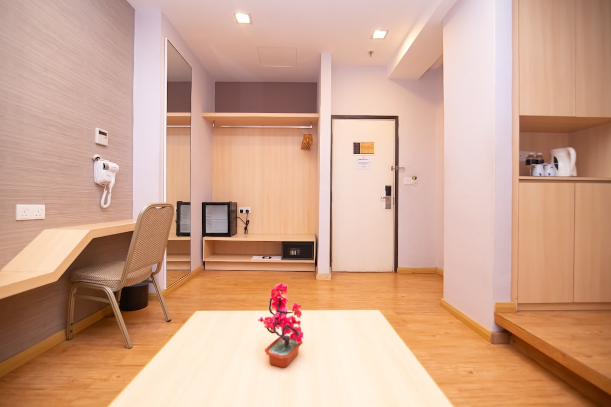 A multifunctional living area is equipped with a wooden table and a single chair. A flat-screen TV and safe are discreetly placed along the wall. The space is complemented by simple shelving and a mirror. A small potted plant adds a touch of color to the table.