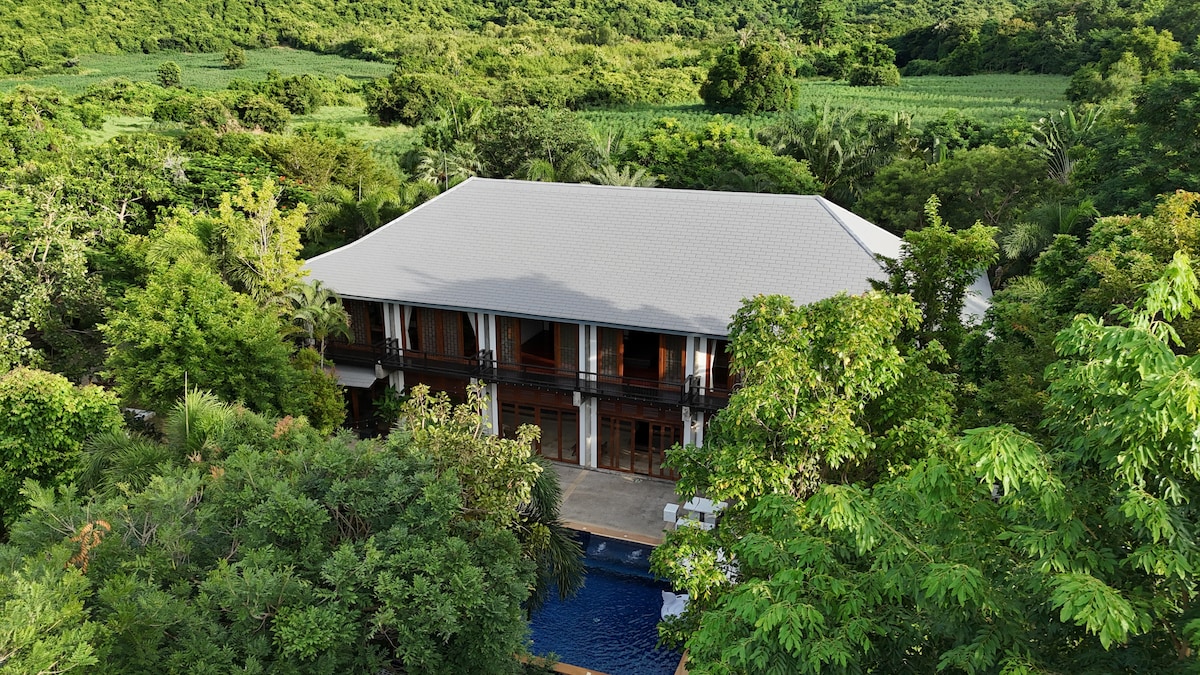 The villa is nestled among lush greenery, showcasing its spacious structure and expansive outdoor area. The inviting pool reflects the surrounding landscape, while large windows offer views of the vibrant natural scenery, providing a serene sense of privacy and tranquility.