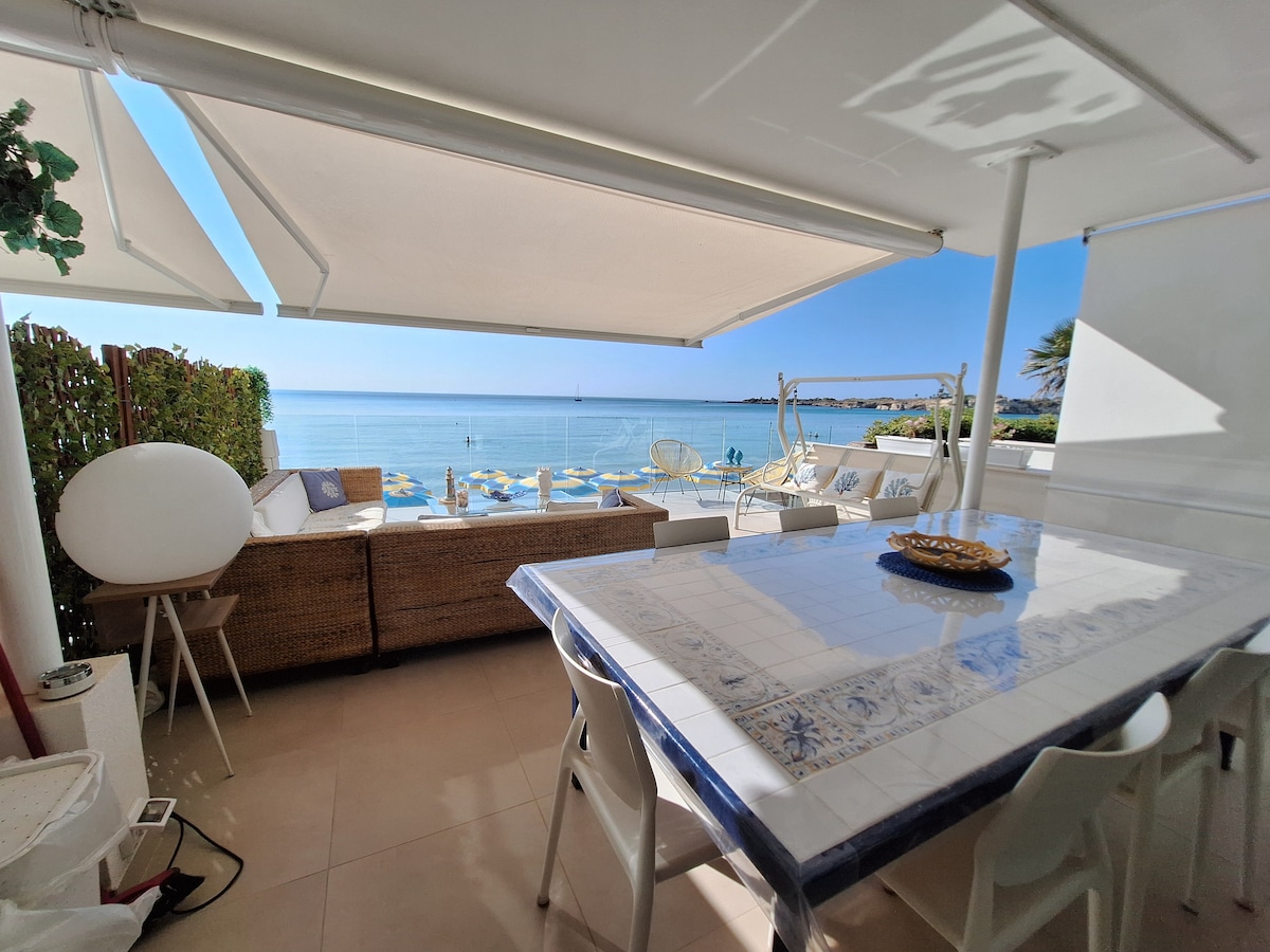 An outdoor dining area is presented with a large table adorned with a decorative centerpiece. Comfortable seating and umbrellas provide shade, with a clear view of the serene beach and calm waters. The space is designed for relaxation and enjoying coastal scenery.