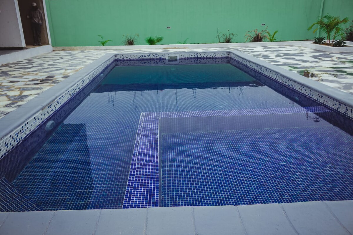A clear swimming pool is framed by tiles in varying patterns. The pool features a unique shape, with steps leading into the water. Lush greenery is visible in the background, contrasting with the light-colored walls surrounding the area.