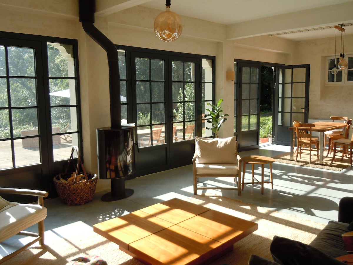A bright living space features large glass doors that invite natural light. Wooden furniture pieces are arranged for comfort, including a cozy armchair and a wooden coffee table. A small plant adds a touch of greenery, while a wood-burning stove provides warmth.