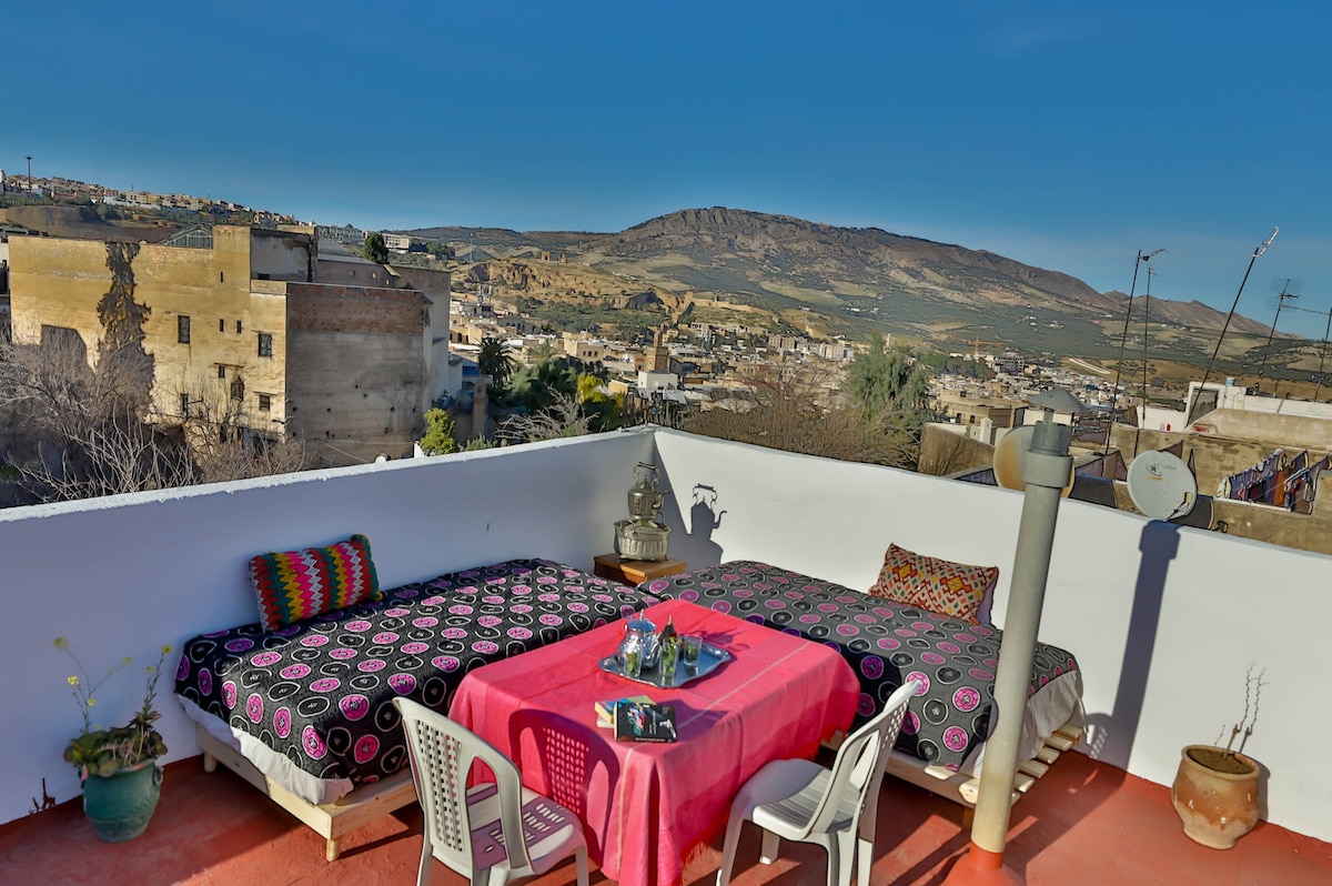 A rooftop terrace features two low beds with colorful beddings and a table set in the center. Surrounding views showcase distant hills and a variety of rooftops. Potted plants are placed along the edge, adding a touch of greenery to the vibrant setting.