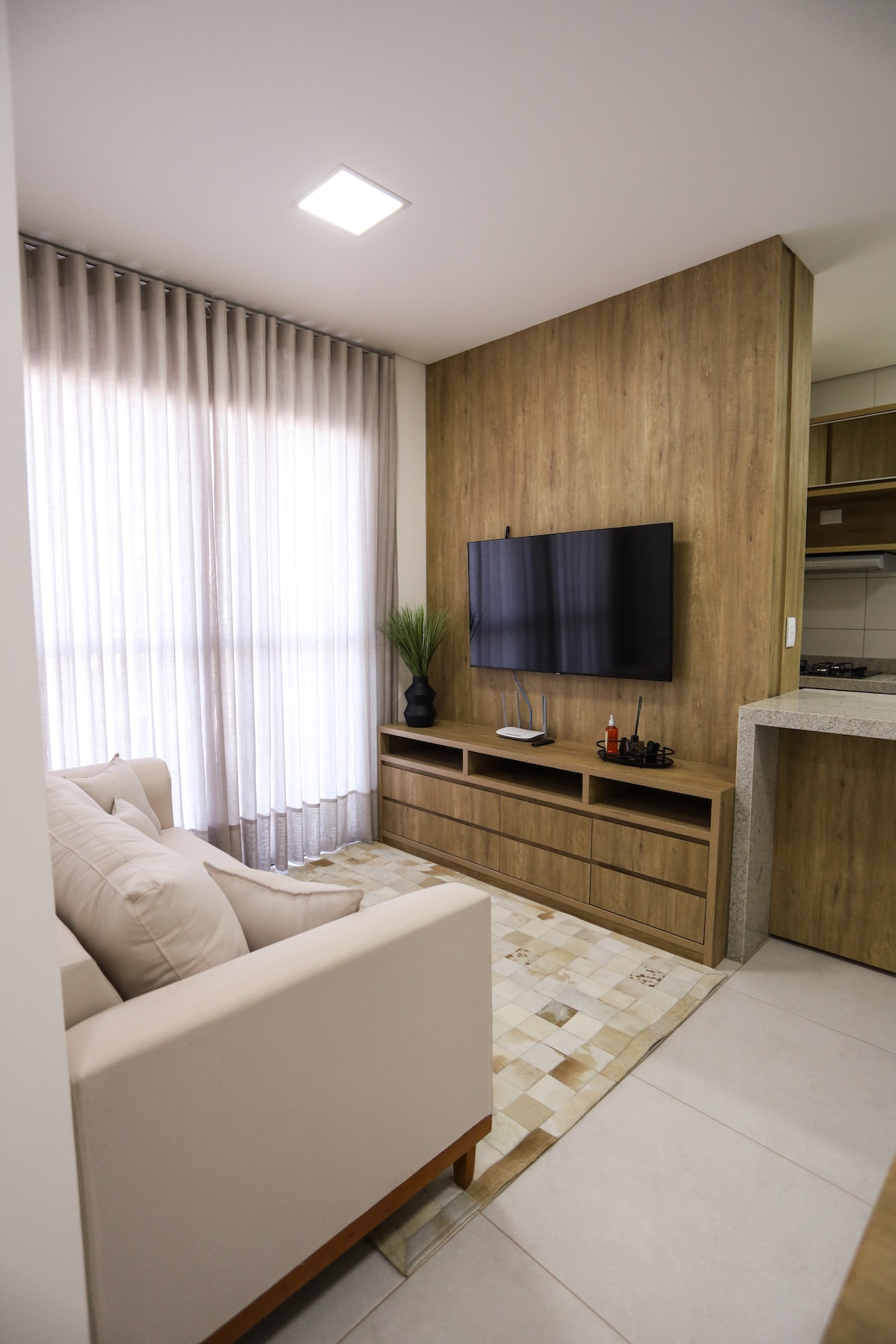 A cozy living area features a light-colored sofa facing a mounted TV, mounted on a wooden wall unit. Sheer curtains allow natural light to filter in, creating a bright atmosphere. A small area rug complements the neutral tones of the room.