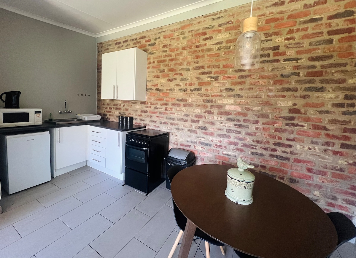 An open plan kitchen is visible, featuring a white refrigerator, microwave, and gas stove set against a brick wall. A round dining table with natural wood legs occupies the center, while a decorative container adds a subtle touch. Ample workspace is provided on the countertop.