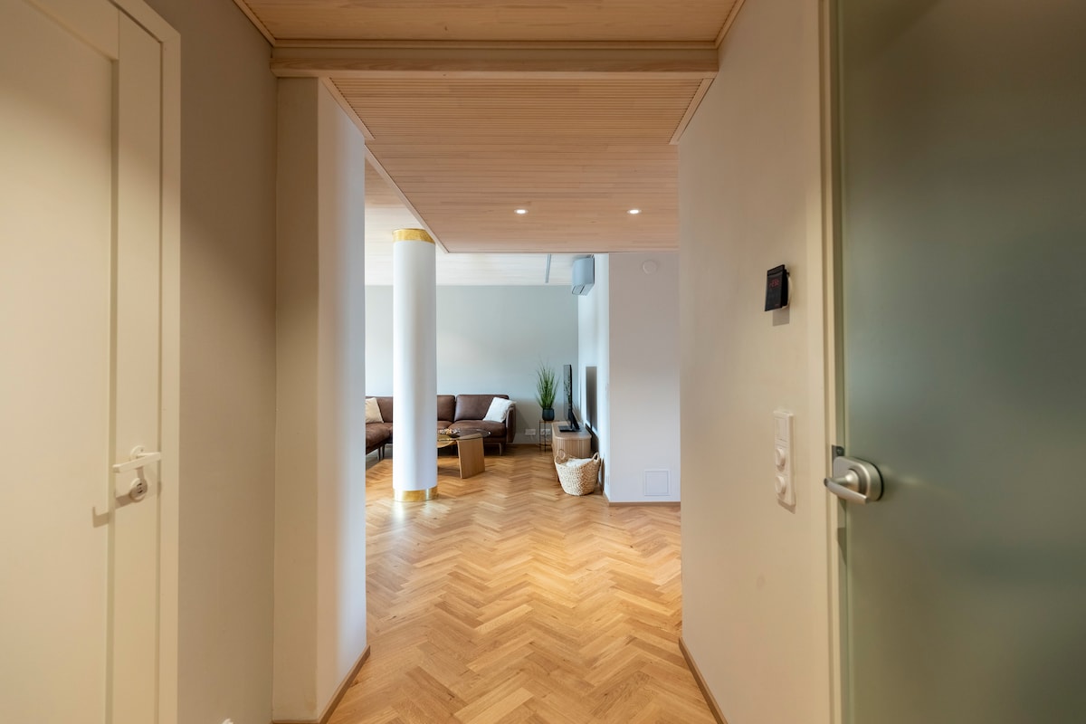 A spacious hallway adorned with herringbone-patterned wooden flooring leads towards a bright living area. Natural light illuminates the space, which is complemented by modern furnishings and a touch of greenery from potted plants set nearby.