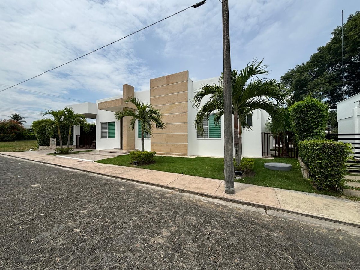 A modern single-story house is displayed with a light-colored façade and stone accents. Lush green landscaping surrounds the entrance, featuring palm trees and neat shrubs. The property is positioned on a quiet street, highlighting a welcoming front yard and a well-maintained driveway.