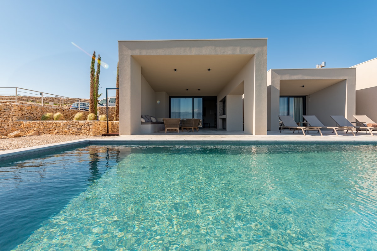 A serene view of the villa's exterior showcases an inviting infinity pool, reflecting the clear blue sky. Sun loungers are arranged beside the pool, providing a spot for relaxation. The minimalist architecture features large openings, enhancing the connection between indoor and outdoor spaces.