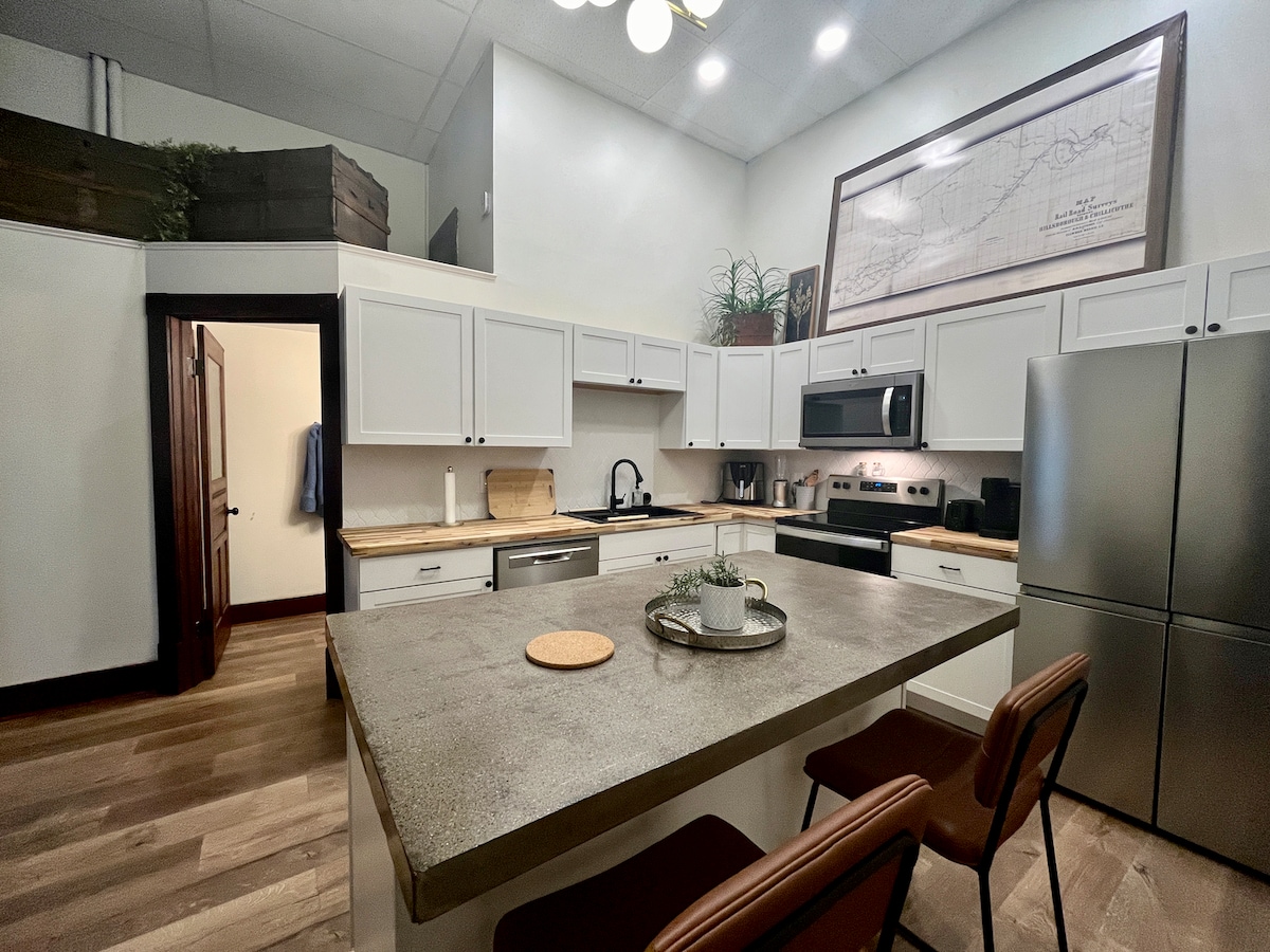 The kitchen features modern appliances, including a stainless steel refrigerator and a sleek microwave. A spacious island with bar seating is positioned at the center, complemented by light wood countertops. Ample cabinet storage and decorative plants enhance the room's inviting design.