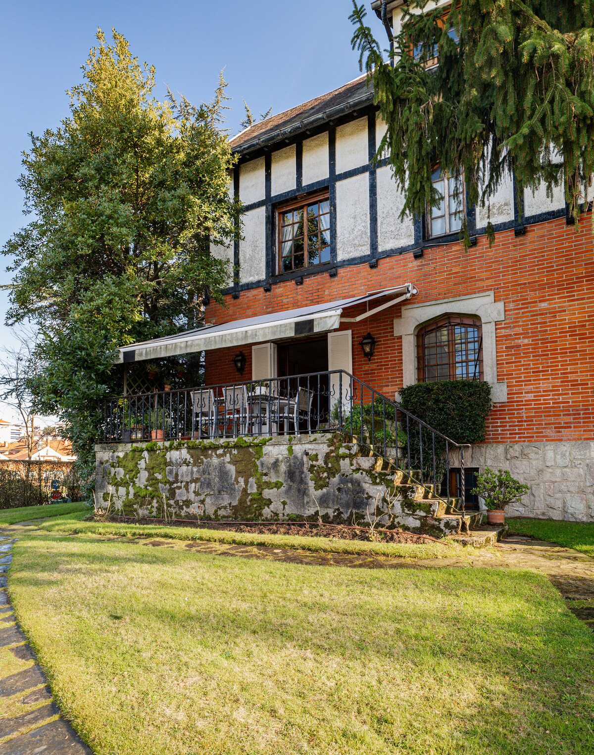 The exterior of the house features a mix of brick and stucco finishes, framed by lush greenery. A spacious terrace with multiple chairs overlooks the manicured lawn, while stone steps provide access to the entryway. Tall trees add to the picturesque surroundings.