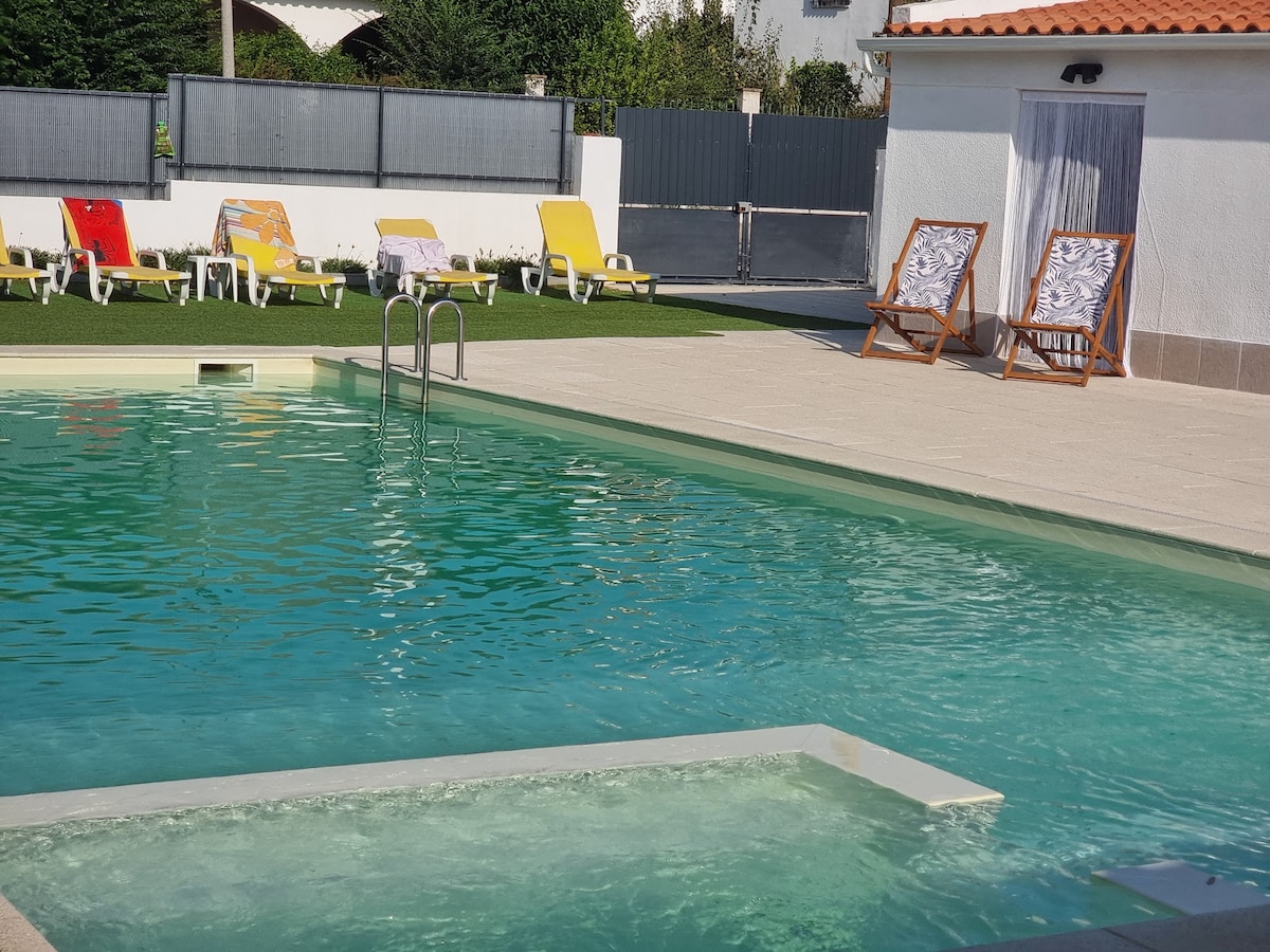 A glistening pool is surrounded by lounge chairs, inviting relaxation under the sun. Two sturdy wooden deck chairs with patterned fabric sit beside the water, enhancing the outdoor setting. Lush green grass frames the area, providing a natural feel to the leisure space.