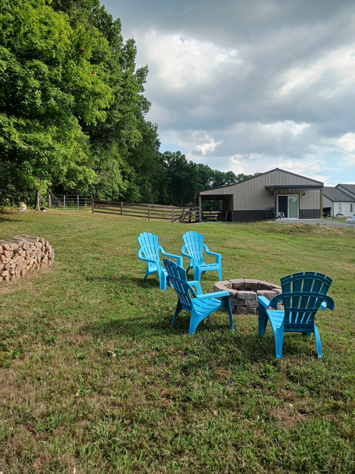 A serene outdoor space features a circular fire pit surrounded by several bright blue Adirondack chairs. Lush greenery and trees provide a natural backdrop, with a structure visible in the distance under a partly cloudy sky. The area is clearly inviting for relaxation and gathering.