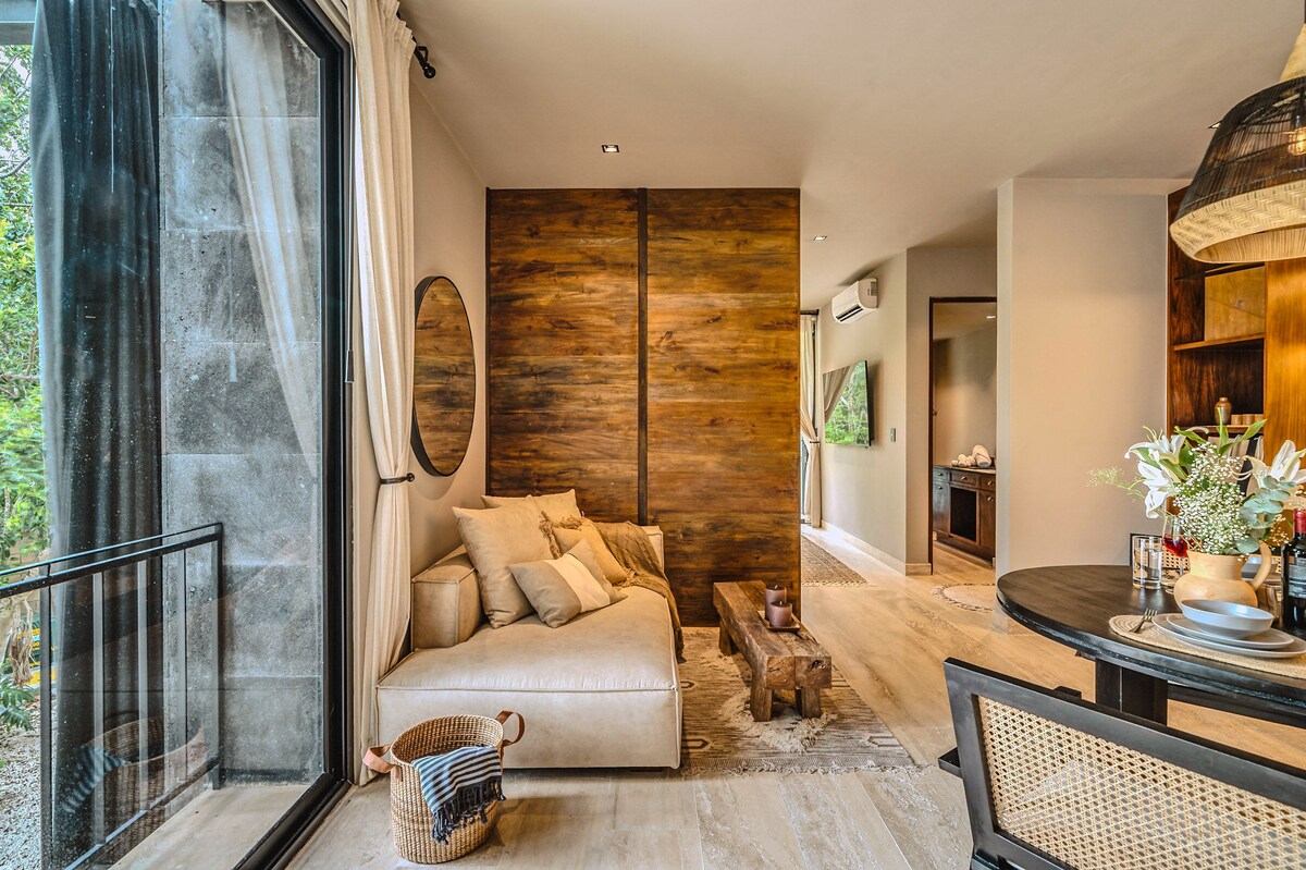 A spacious living area is featured with a contemporary sofa and a round mirror on the wall. Natural wood accents create warmth, while the open layout leads to a well-equipped kitchen area visible in the background. Large glass doors provide access to outdoor views.