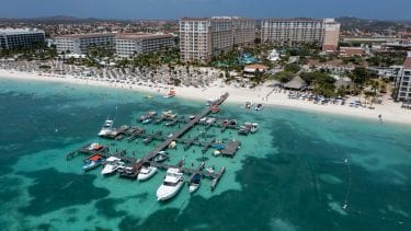 An aerial view reveals a vibrant beach scene featuring a wooden dock extending into clear turquoise waters. Multiple boats are moored alongside the dock, while sandy beach chairs are arranged along the shore. Resort buildings are visible in the background, surrounded by palm trees.