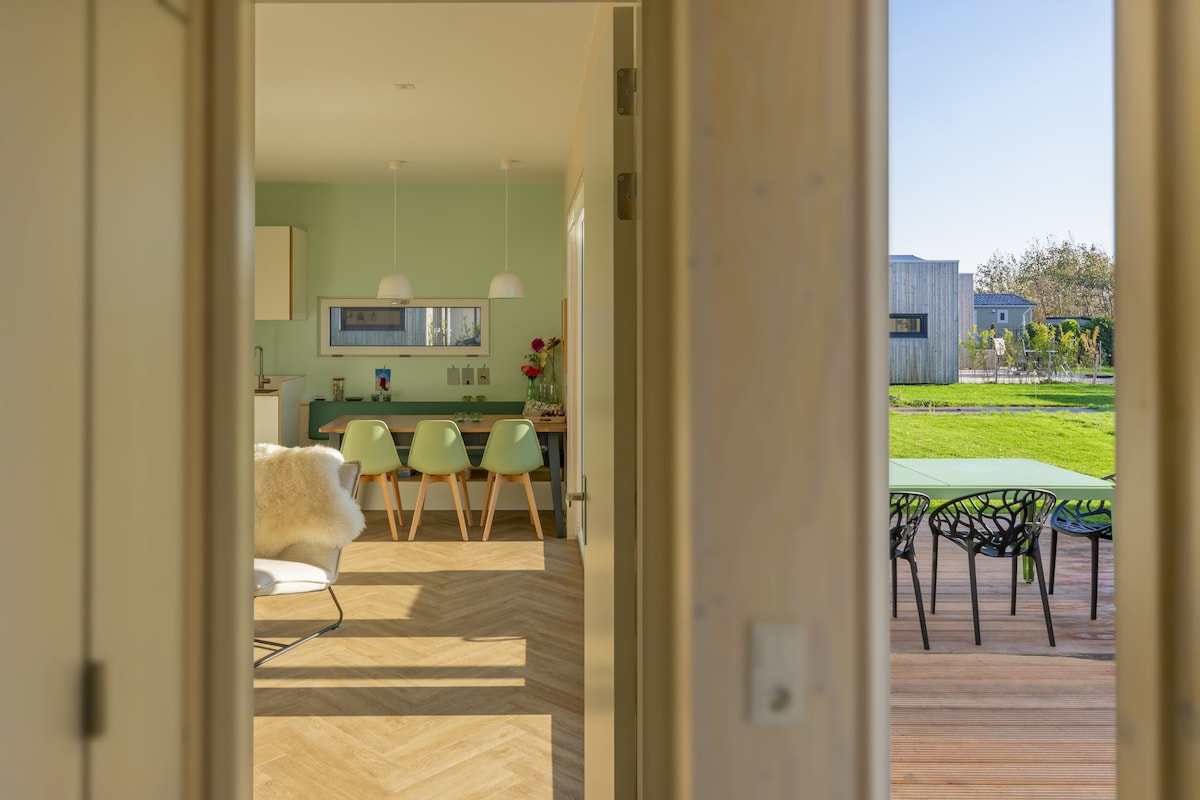 A doorway frames a view into a modern kitchen and dining area, featuring soft green walls and a light wooden floor. Beyond, a dining table is paired with pale chairs, while an outdoor space with green tables and black chairs is visible through large windows.