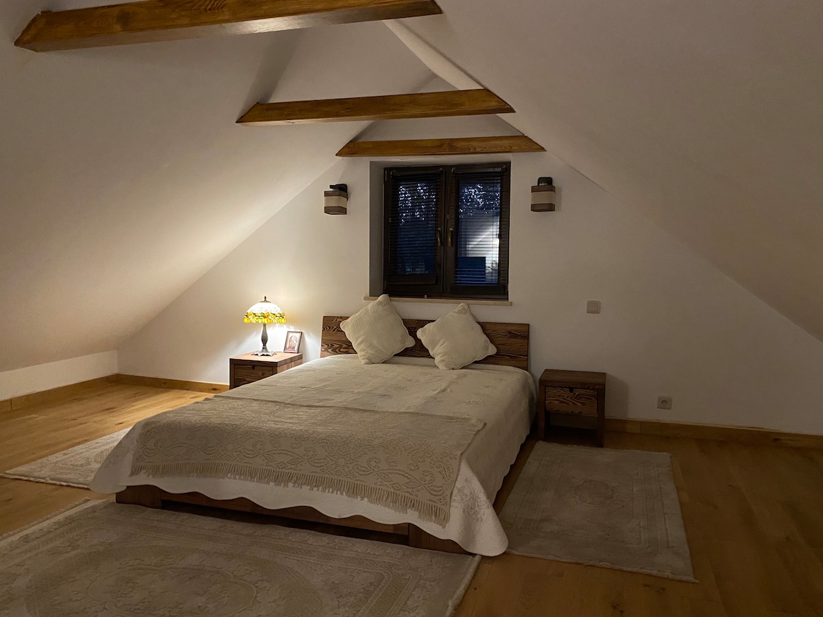 A spacious bedroom features a large bed with light bedding and decorative pillows. Two bedside tables are positioned on either side of the bed. Windows with wooden frames allow soft light to enter, while wooden beams add to the room's architectural charm.