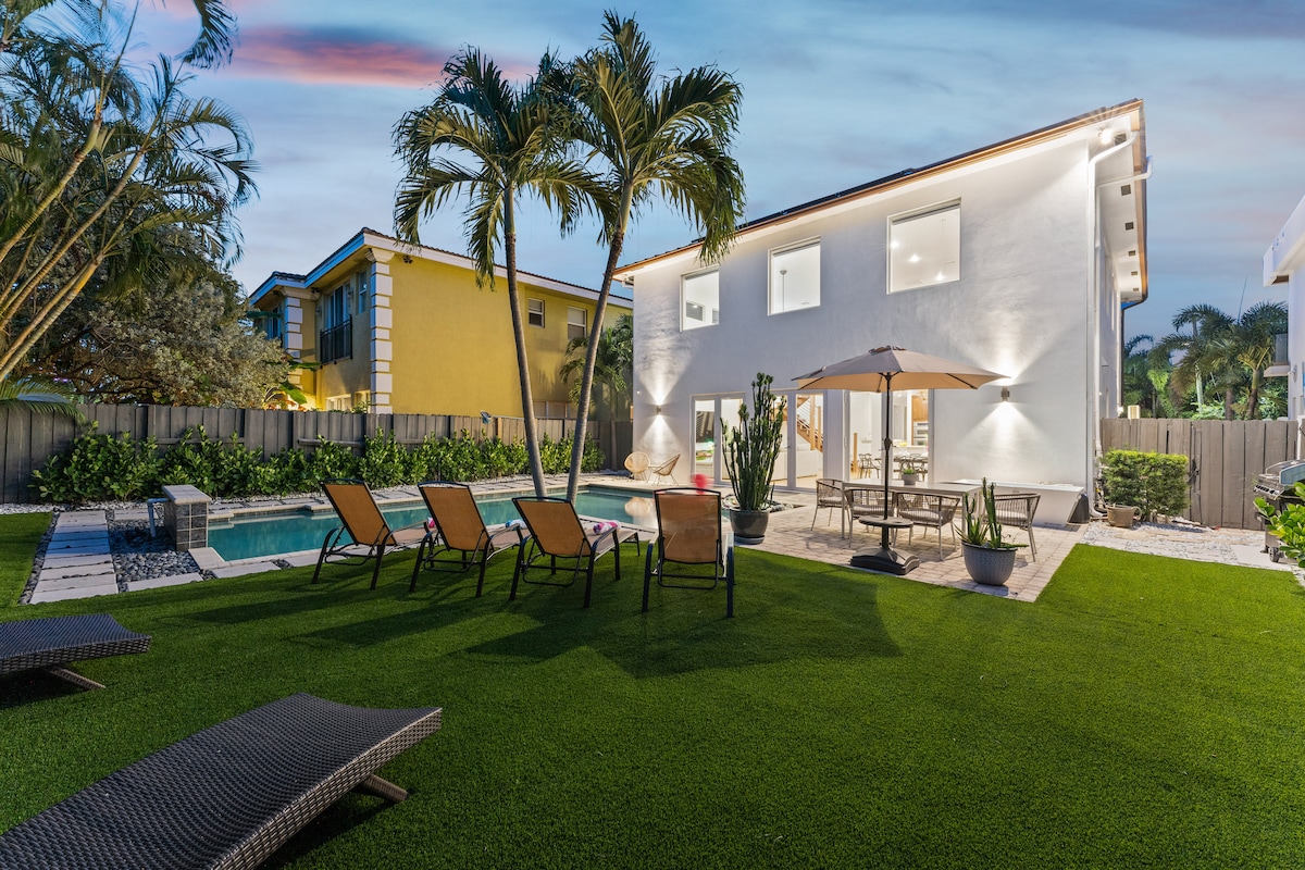 The backyard features a well-manicured lawn surrounding a heated pool, with lounge chairs positioned nearby. A dining table under an umbrella sits adjacent to the house, providing an inviting space for outdoor meals. Tropical plants enhance the serene atmosphere of the retreat.