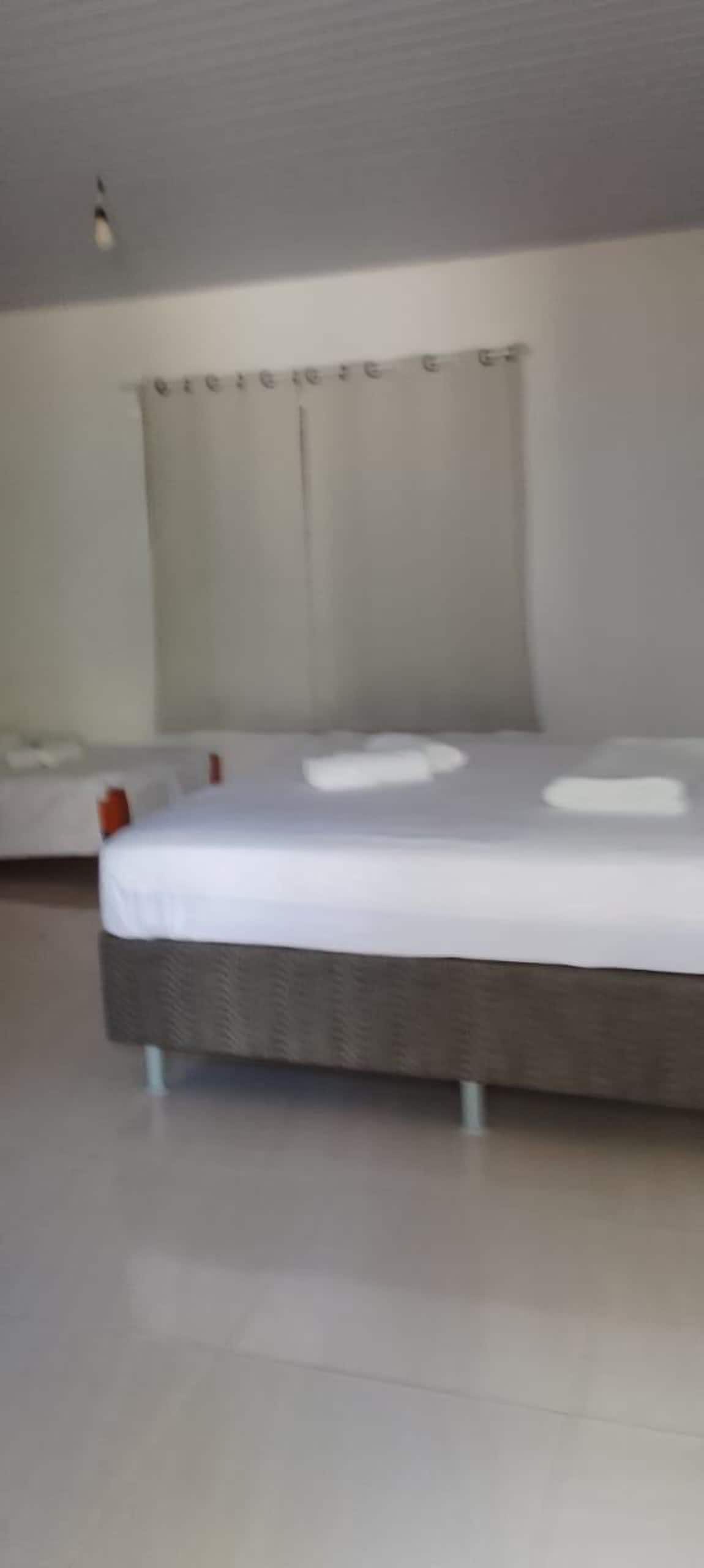 A spacious sleeping area features a large bed with fresh linens and two decorative pillows. A smaller bed is positioned nearby, complemented by soft, neutral tones and natural light filtering through partially drawn curtains.