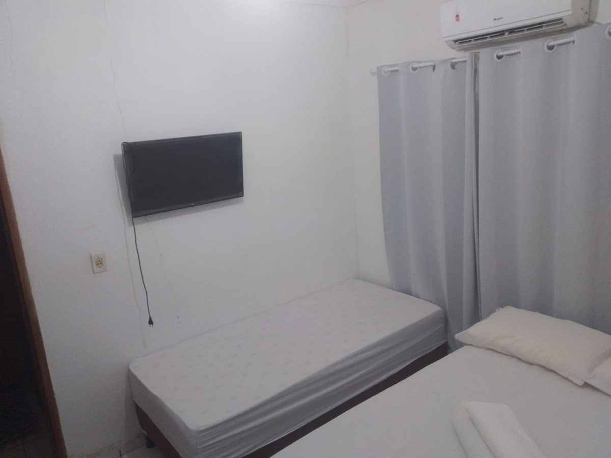 A simple sleeping area features two beds, one unmade twin bed and a made double bed. A wall-mounted television is positioned in front of the beds. Light-colored curtains frame a window, allowing natural light to fill the space.