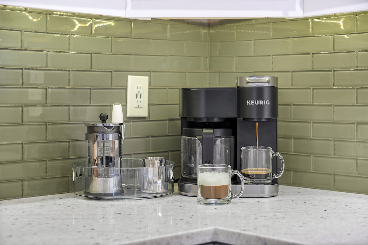 A well-equipped coffee station is situated on a sleek countertop. A Keurig machine and French press are displayed alongside glass mugs filled with coffee. The backsplash features green glass tiles, enhancing the modern aesthetic of the kitchen.
