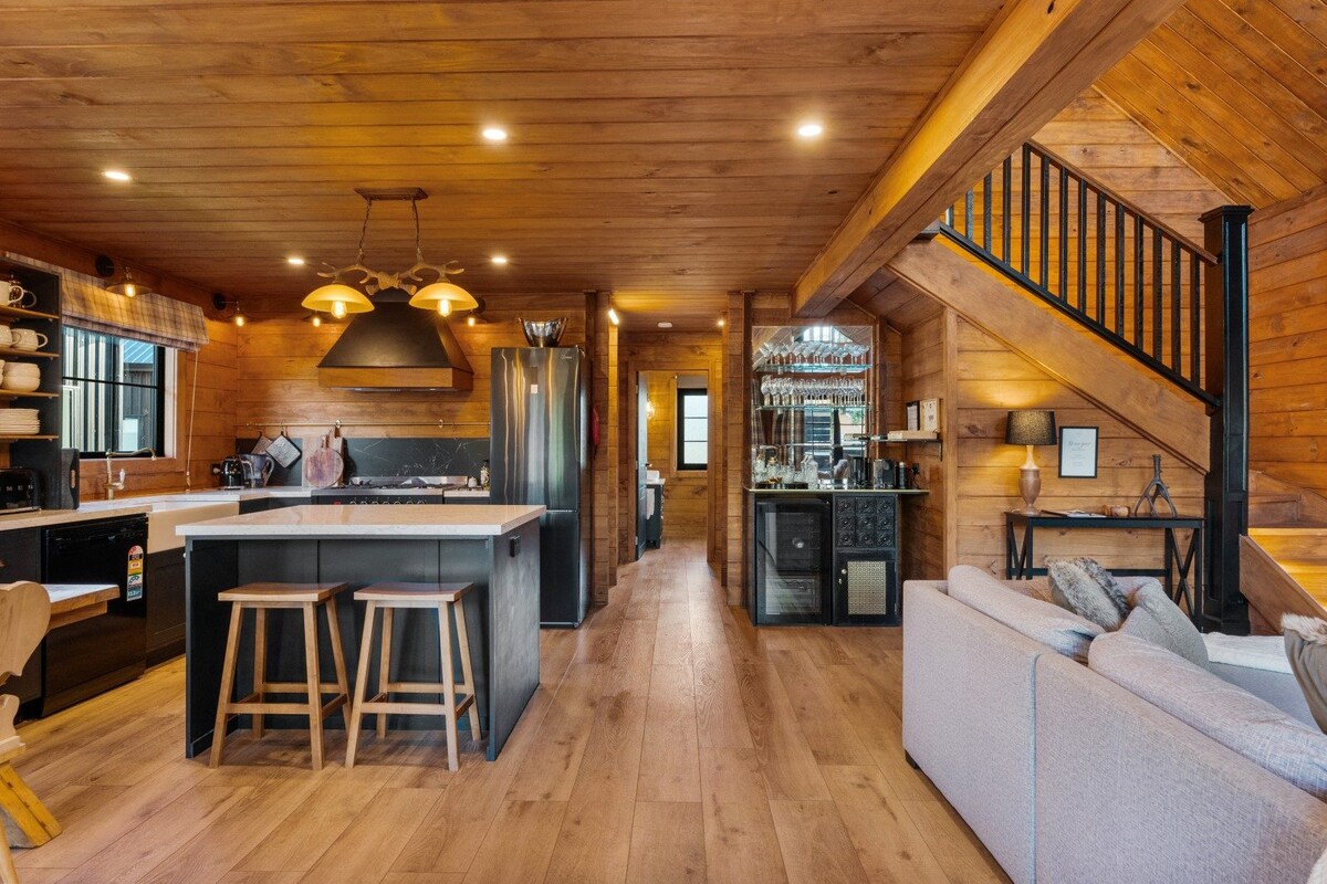 A spacious open-plan interior features wooden paneling and warm lighting. The kitchen is equipped with modern appliances, including a sleek black refrigerator. A cozy seating area is set beside a staircase leading to the upper level, while two wooden stools are positioned at a kitchen island.