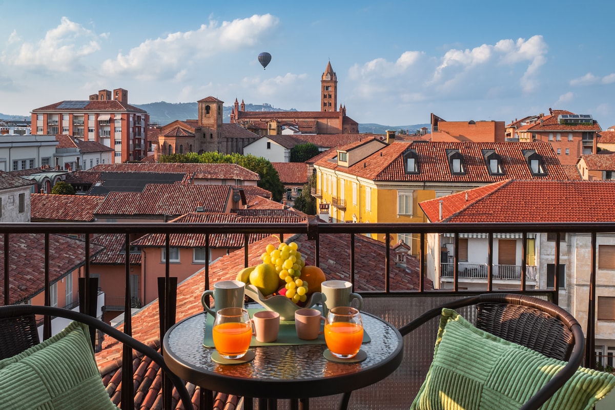 A balcony offers a stunning view of Alba's rooftops and the distant hills, with a colorful hot air balloon drifting in the sky. A table is set with fresh fruit, two glasses of orange juice, and several mugs, inviting relaxation and enjoyment of the scenery.