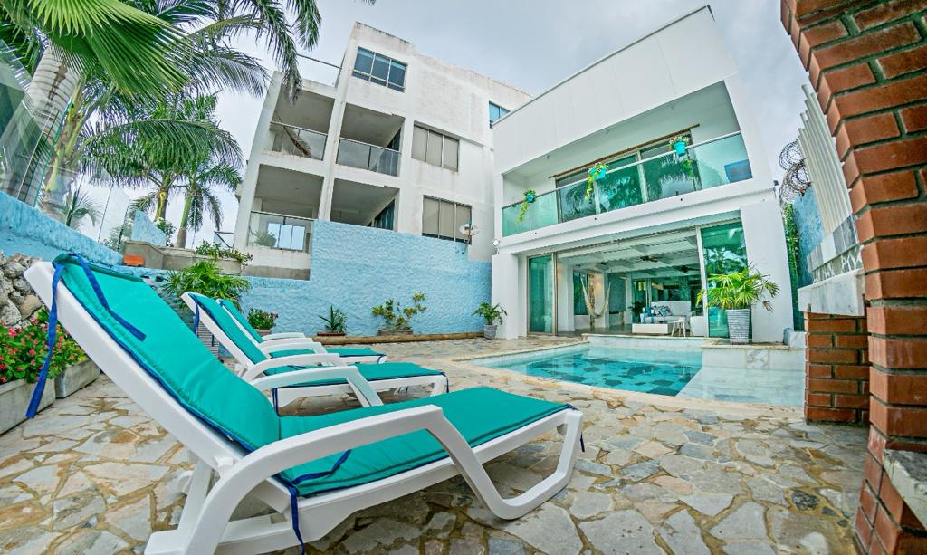 A tranquil outdoor area is highlighted by a pool surrounded by natural stone tiles. Lounge chairs with teal cushions are positioned nearby. The facade of a modern building is visible in the background, alongside lush greenery and palm trees, enhancing the relaxing vibe.