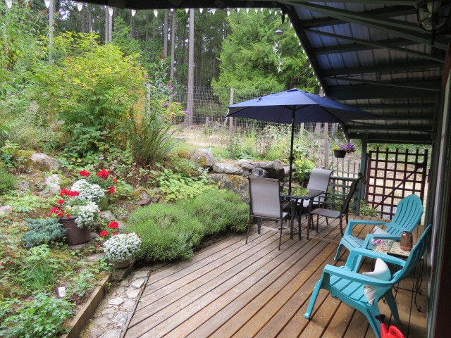 A covered patio area features a table with four chairs and two blue lounge chairs, set on a wooden deck. A garden with blooming flowers and lush greenery is visible in the background, along with a stone wall enhancing the natural setting.