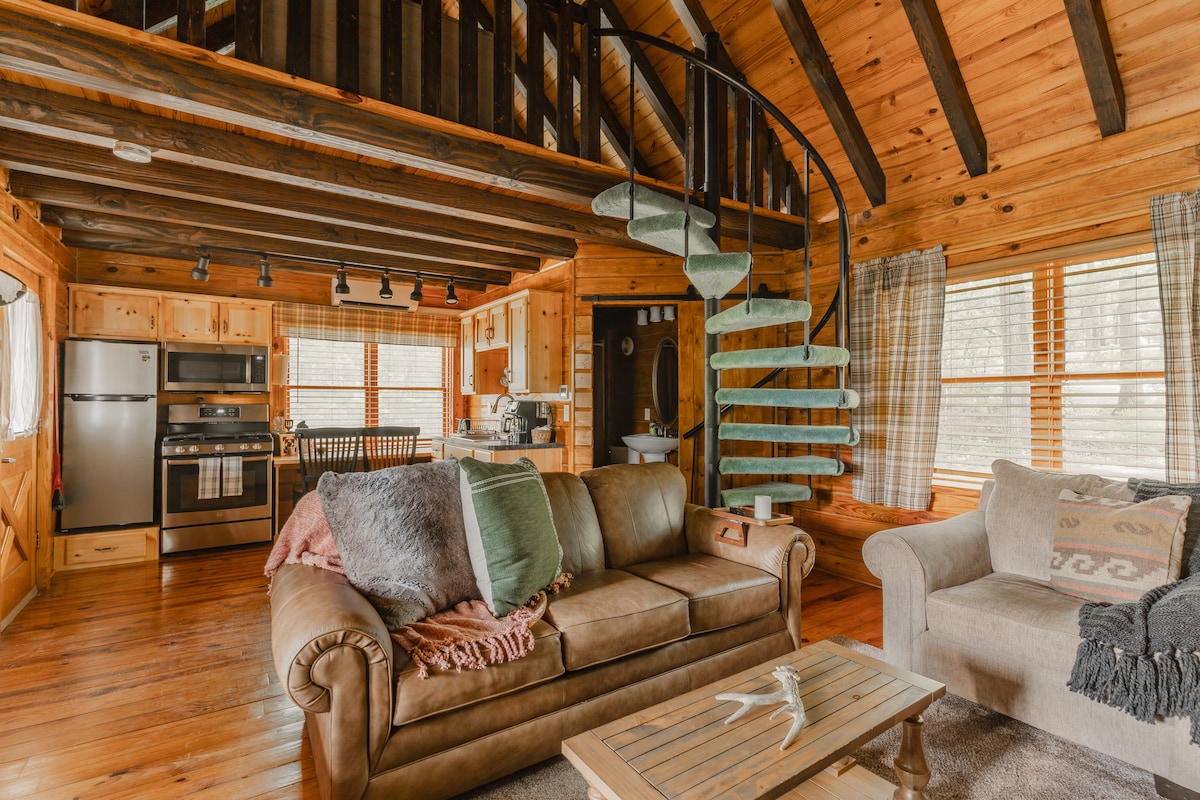 A spacious living area is highlighted by exposed wooden beams and rich hardwood floors. A comfortable leather sofa is adorned with soft blankets and pillows. Adjacent, a well-equipped kitchen can be seen, complemented by a spiral staircase leading to a loft area.
