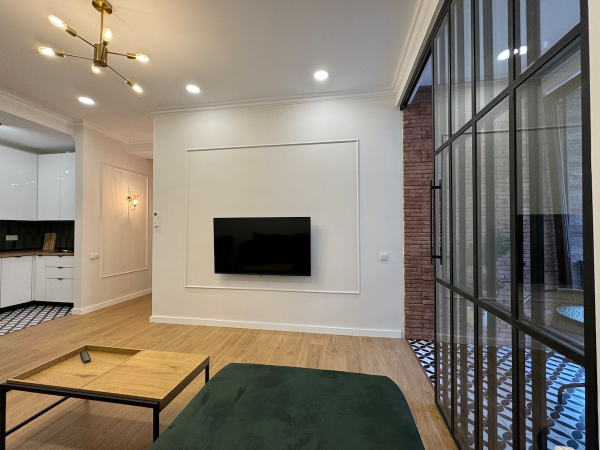 A modern living space showcases a flat-screen television mounted on a feature wall. A low coffee table rests on a wooden floor, while large glass doors provide a view of the patio. Soft lighting is provided by a stylish ceiling fixture.
