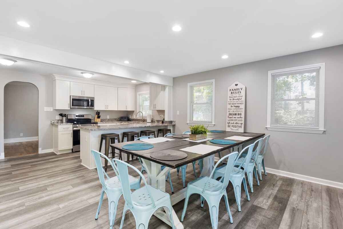 A spacious dining area features a large wooden table surrounded by light blue chairs. Bright, natural light enters through multiple windows. A modern kitchen with white cabinetry and stainless steel appliances is visible in the background, creating an open and inviting space for gatherings.