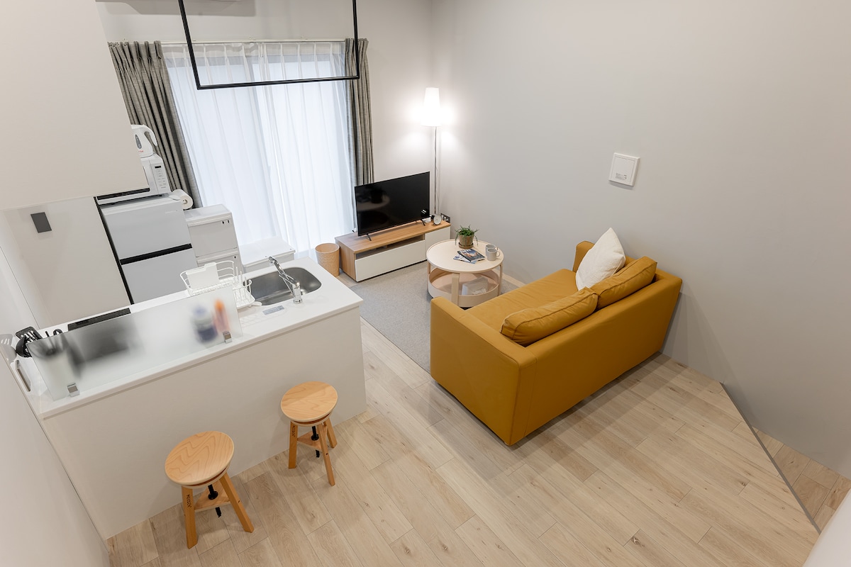 A modern living space is presented, featuring a cozy yellow sofa facing a television. Light streams in through large windows, complemented by soft curtains. A dining area with two stools and a minimalist kitchen are visible, creating an inviting atmosphere for relaxation and socializing.