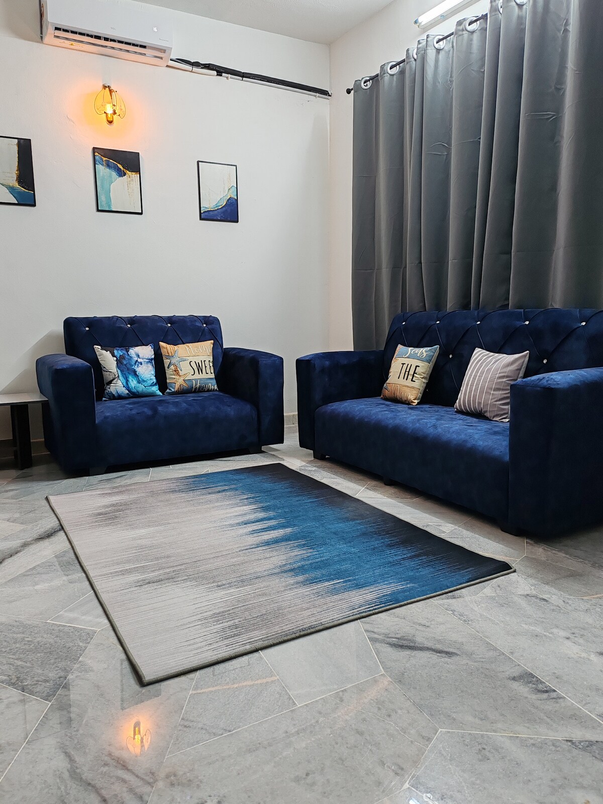 A minimalist living area is presented with two blue velvet sofas, adorned with decorative cushions. A soft area rug with an ombre design sits on the polished marble floor. Soft lighting from a wall sconce complements the gray curtains that frame the window.