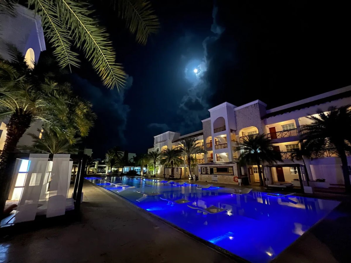 The image showcases a beautifully illuminated pool area at night, surrounded by lush palm trees. The water reflects the moonlight, creating a calming blue hue. The architecture features a modern design, with spacious balconies overlooking the pool, enhancing the tranquil evening atmosphere.