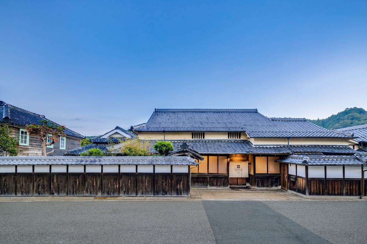 The exterior of a traditional wooden structure is displayed against a deep blue sky, showcasing a distinct sloped roof and intricate wooden detailing. The property is bordered by well-maintained paths and features greenery and rustic fencing, emphasizing its historical charm.