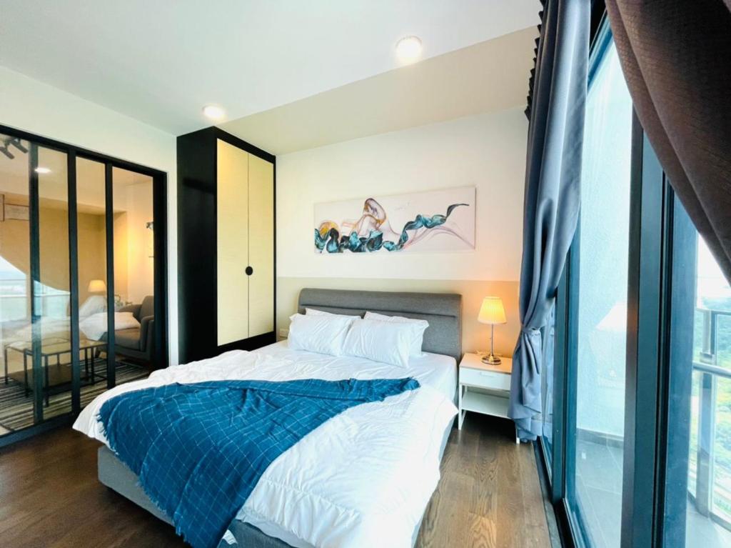 A cozy bedroom features a spacious bed covered with crisp white bedding and a dark blue throw. A bedside table with a lamp provides soft lighting, while large windows allow natural light to fill the room. Stylish artwork adorns the wall, enhancing the ambiance.