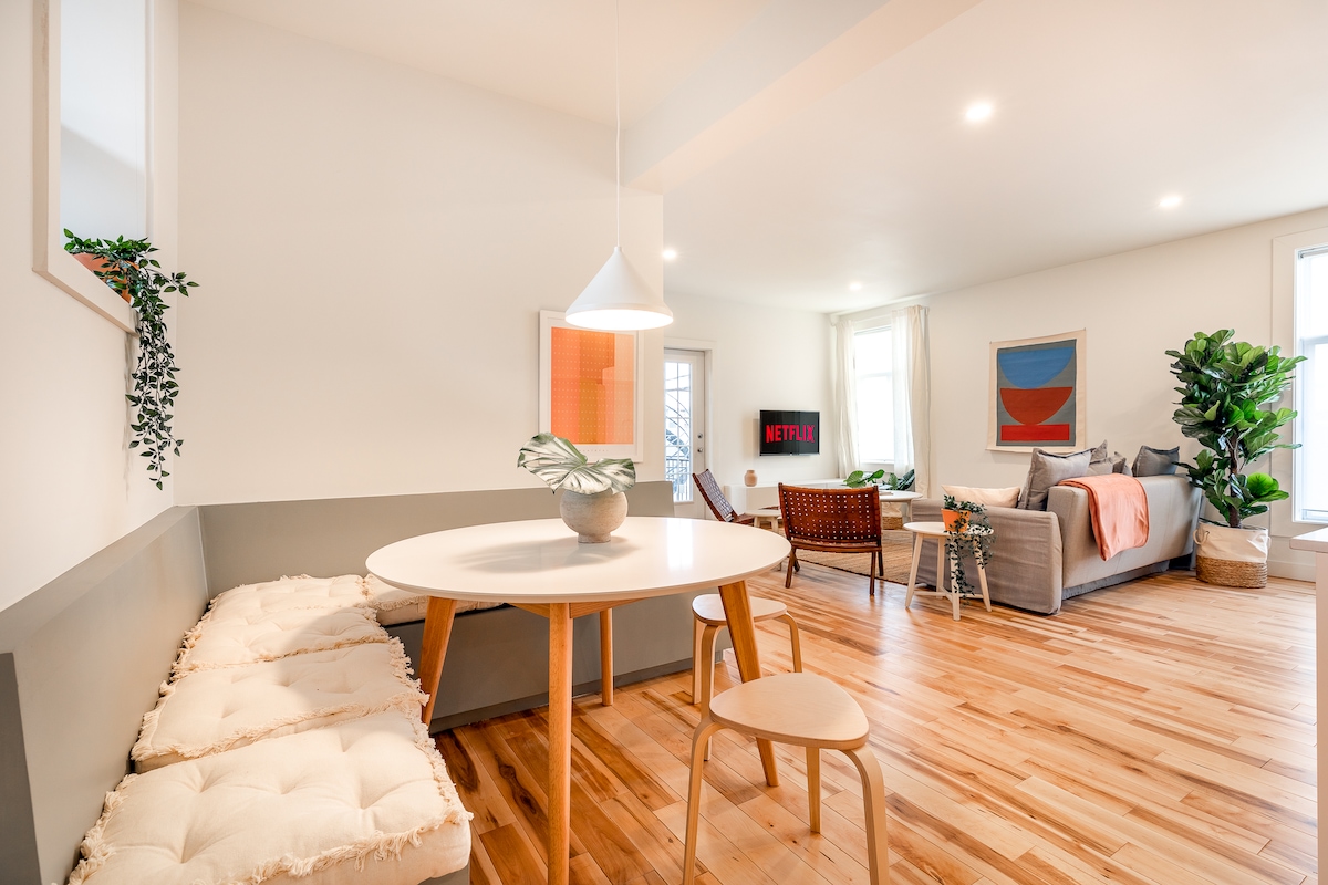 A modern living space features a round dining table with a plush seating area alongside. A comfortable sofa is complemented by decorative pillows, while a flat-screen television is mounted on the wall. Natural light fills the area, highlighting wooden flooring and indoor plants.