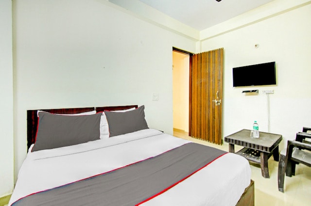 A bright room features a spacious bed with a white and gray duvet, complemented by matching pillows. A small table and two chairs are positioned nearby. A wall-mounted TV is visible, along with a wooden door leading to an adjoining area.