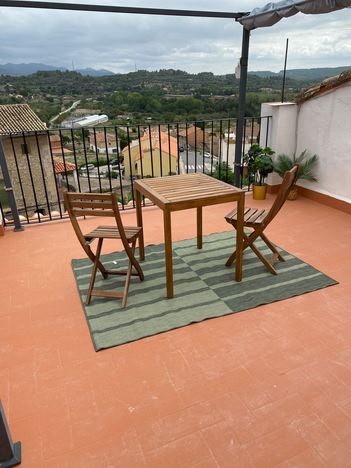 An outdoor terrace is featured, furnished with a wooden table and two folding chairs. A green area rug lies beneath the table, and a potted plant adds a touch of greenery. Scenic views of the valley and nearby hills are visible in the background.