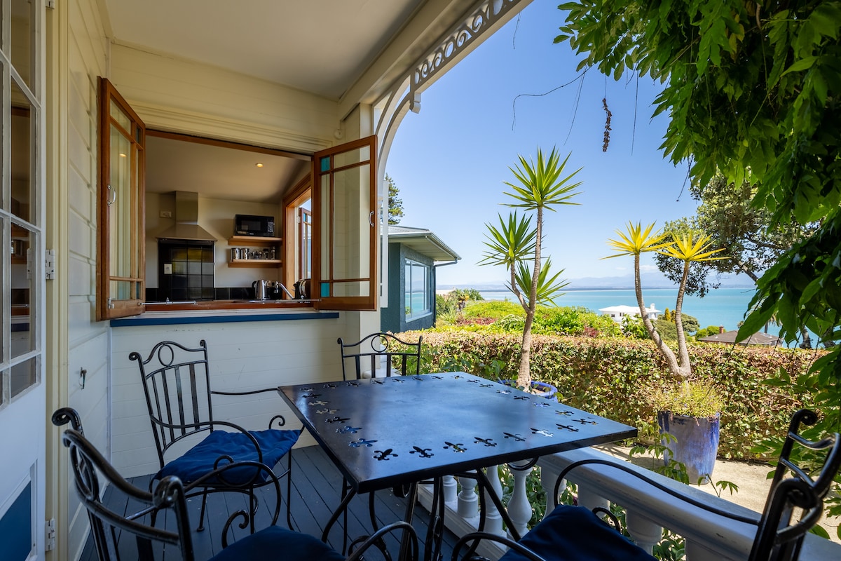 An outdoor seating area features a wrought iron table surrounded by four chairs, set against a backdrop of vibrant greenery and ocean views. Light filters through open windows into the kitchen area, creating a seamless connection between indoor and outdoor spaces.