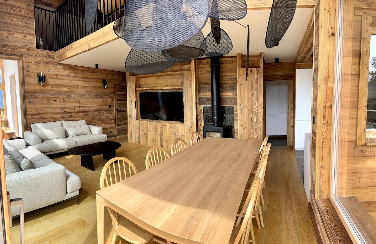 A spacious living area is characterized by natural wooden walls and stylish furnishings. A large dining table is surrounded by light-colored chairs, while a comfortable seating arrangement is positioned nearby. A television is mounted on the wall, and a wood stove adds warmth to the space.
